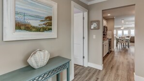 A hallway with a painting of a beach scene, a white decorative item on a console table, and a view into a dining area with a wooden floor. - Oceanfront Beach House with Spacious Backyard by RedAwning (Fernandina Beach)