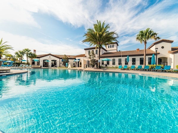 A swimming pool with lounge chairs and umbrellas, surrounded by palm trees and buildings with a Mediterranean architectural style.