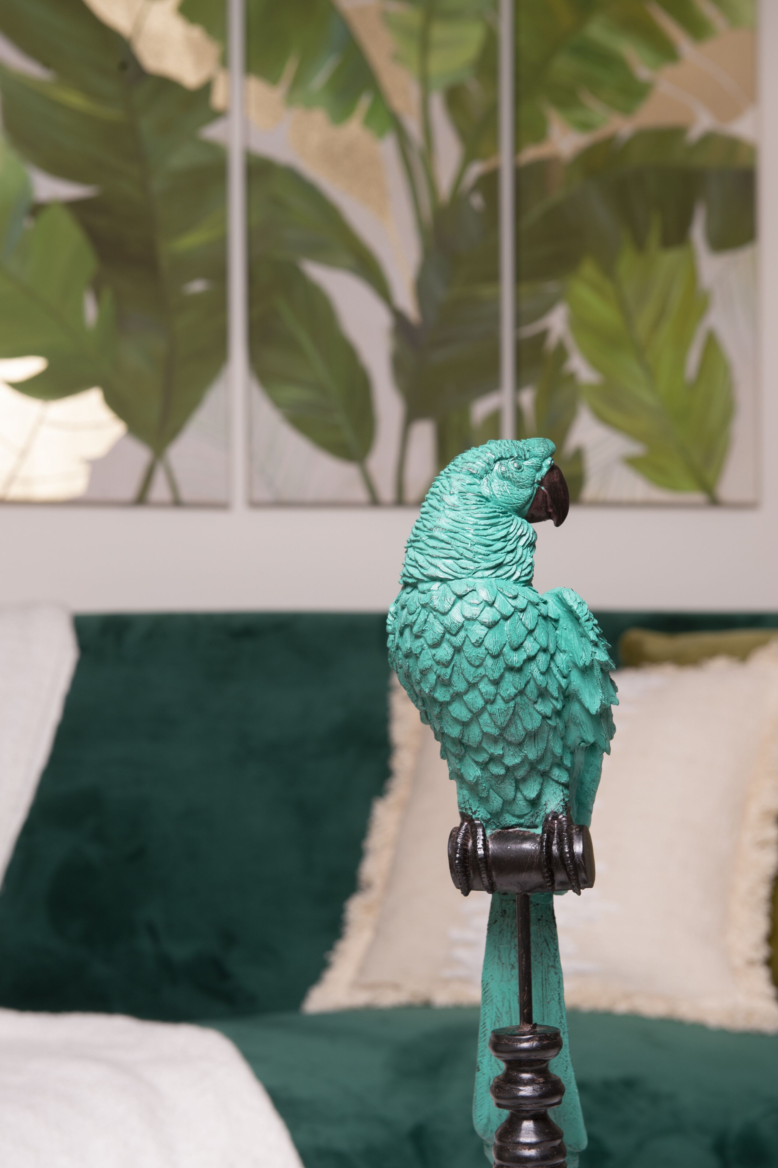 Un perroquet était perché sur un support dans une pièce où se trouvait un couvre-lit à motifs verts et blancs, et une grande plante en arrière-plan.