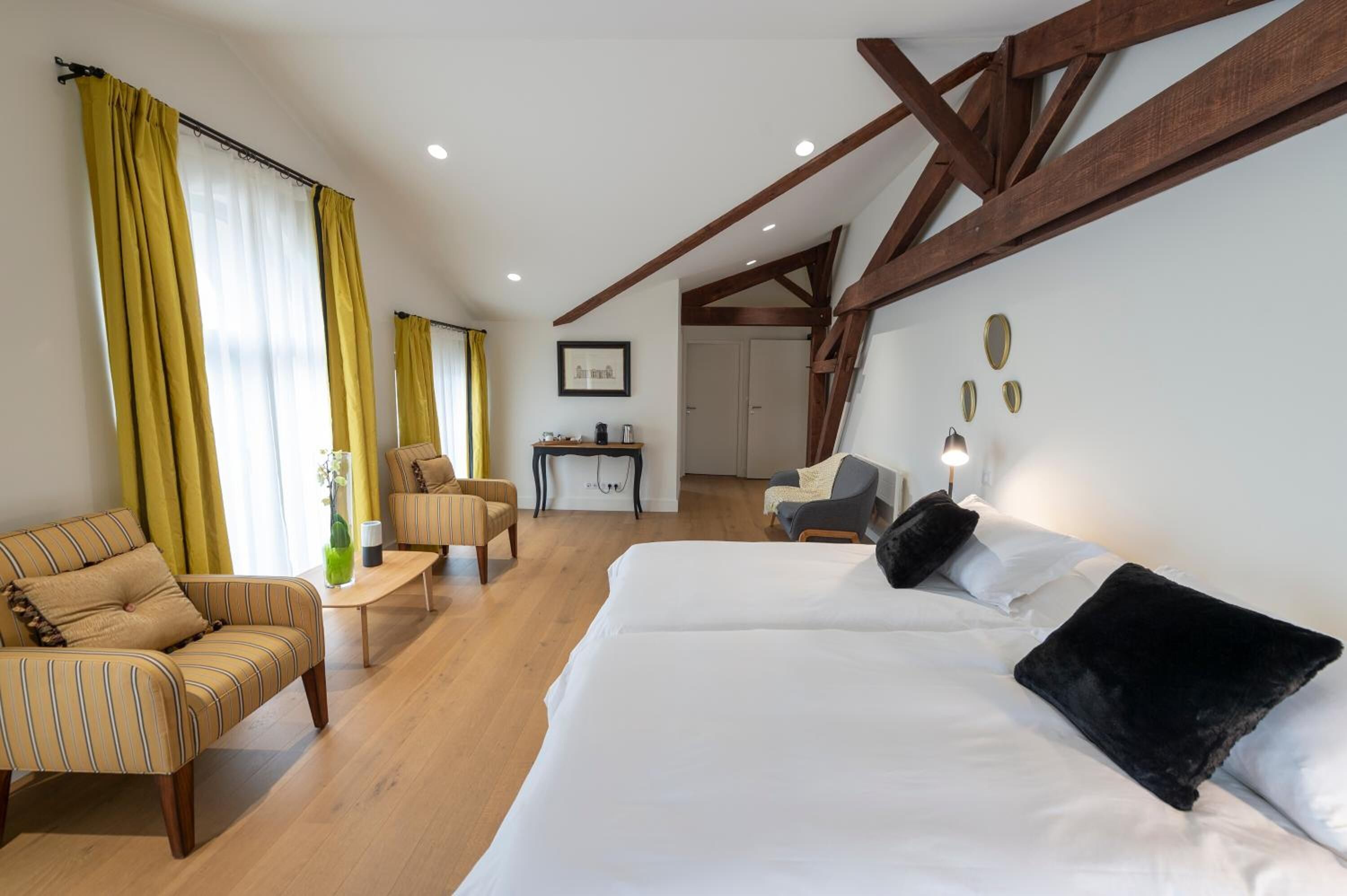 Habitación de hotel con una cama grande, un sofá, un escritorio y una silla. Se ve una ventana con cortinas, techo de madera y una pared con placas decorativas.