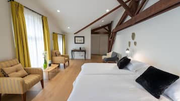 Habitación de hotel con una cama grande, un sofá, un escritorio y una silla. Se ve una ventana con cortinas, techo de madera y una pared con placas decorativas.
