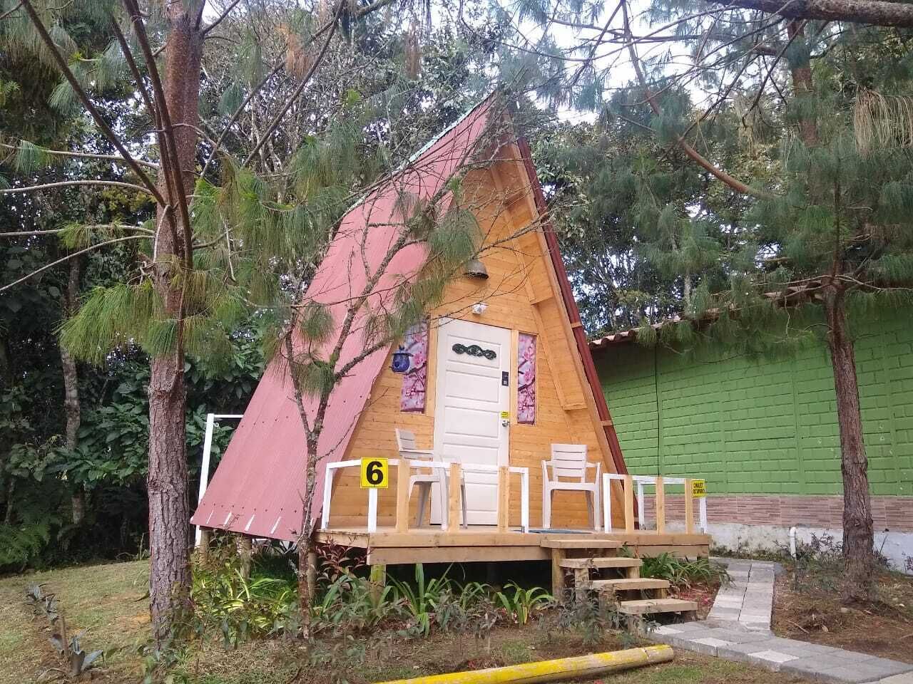 Estrutura de madeira triangular com porta e duas cadeiras na frente, cercada por árvores.