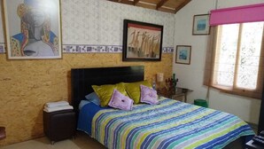 Ein Schlafzimmer mit Bett, Kissen, einem Nachttisch, einem Fenster mit Jalousien und Wandbildern.
