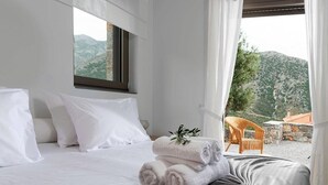 A hotel room with a bed, white pillows, a view of mountains, and a wicker chair outside.