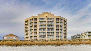 Un hotel a più piani con tetto piatto e balconi, situato su una spiaggia sabbiosa con dune in primo piano.