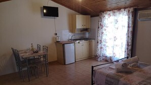 A compact room with a bed, a small kitchenette, a dining table with chairs, and a TV mounted on the wall. - Ocean Front Studios (Kefalonia)