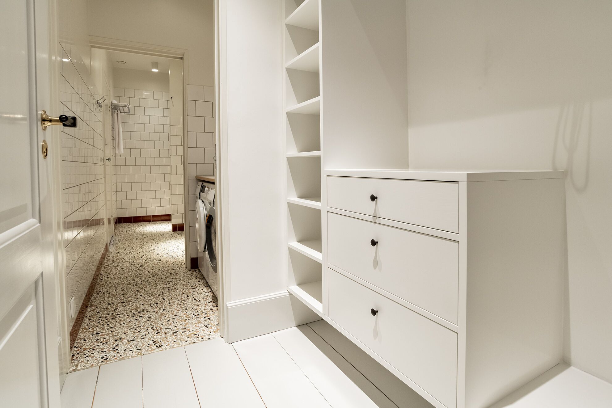 A clean, modern bathroom with white tiles, a built-in washing machine, and a white vanity with drawers.