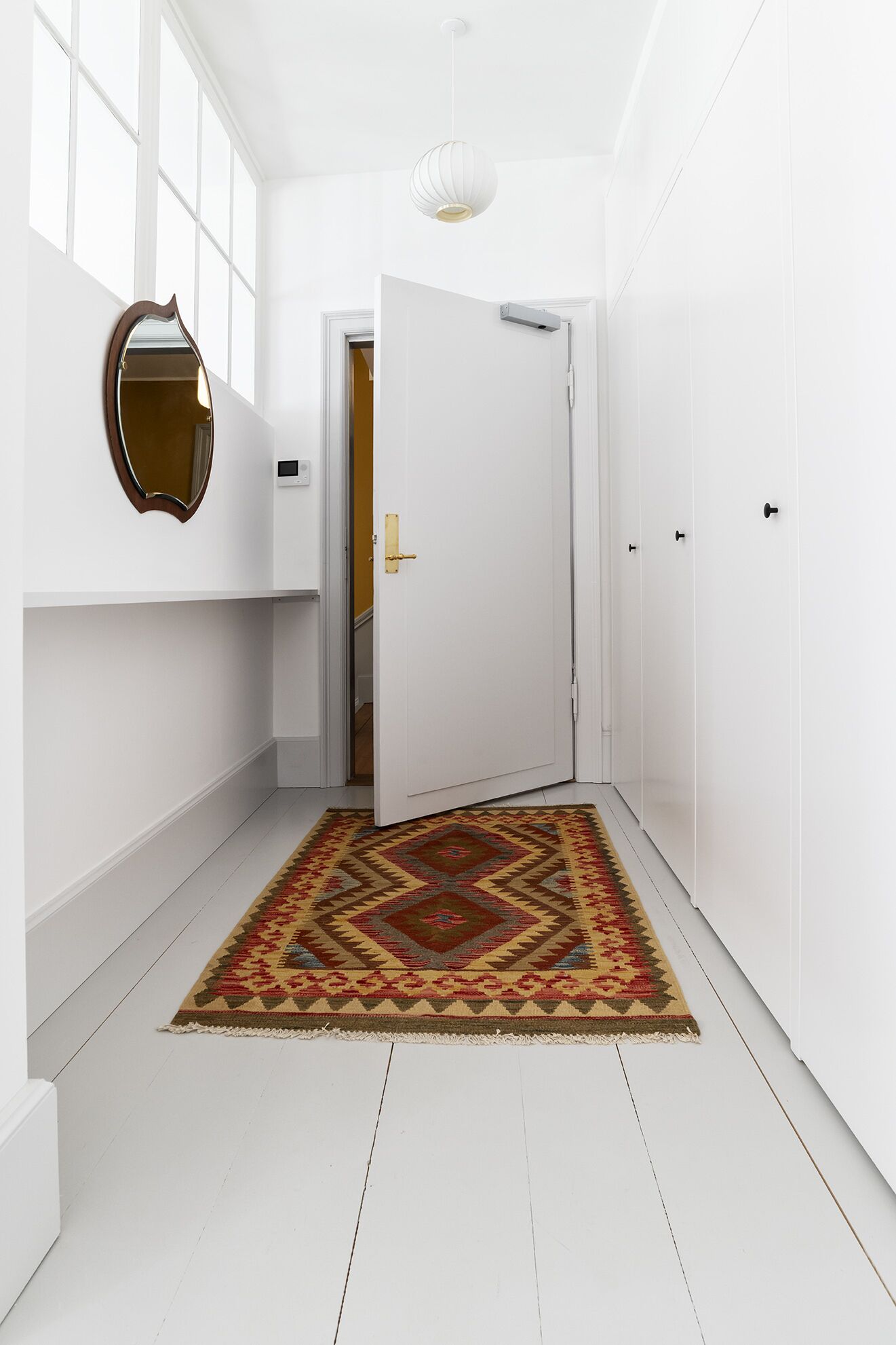 Un pasillo con paredes blancas, un tapete estampado, un espejo y una puerta que da a otra habitación.
