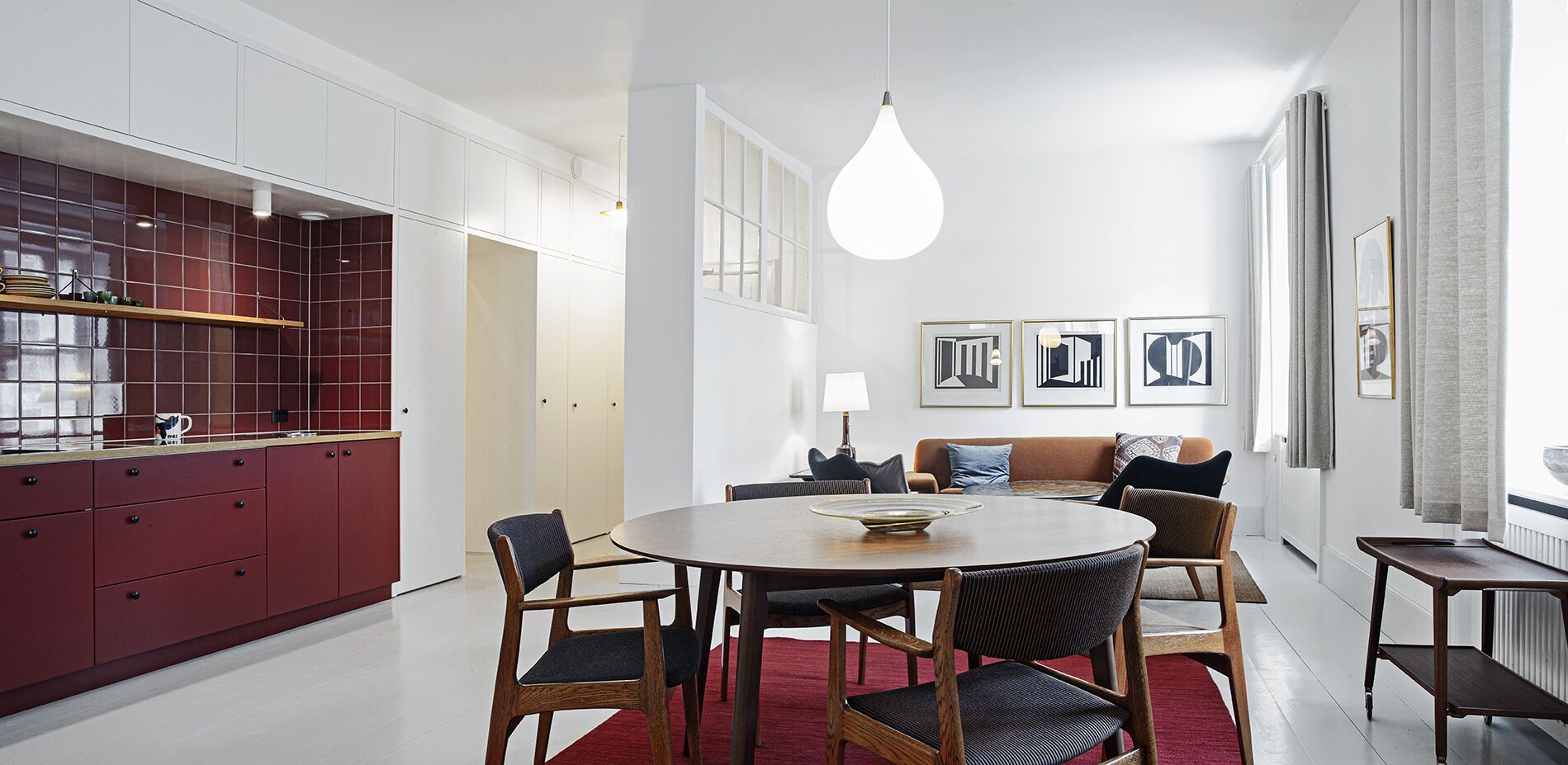 Une cuisine moderne avec des meubles de rangement rouges, un coin repas avec une table ronde et des chaises, et un salon avec un canapé et des œuvres d’art encadrées.