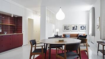 A modern kitchen with red cabinets, a dining area with a round table and chairs, and a living room with a sofa and framed artwork.