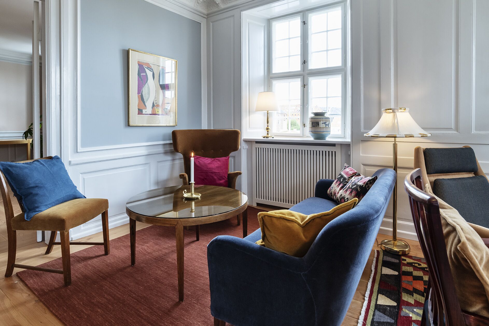 A living room with a round coffee table, two armchairs, a sofa, a radiator, and a window with a view.