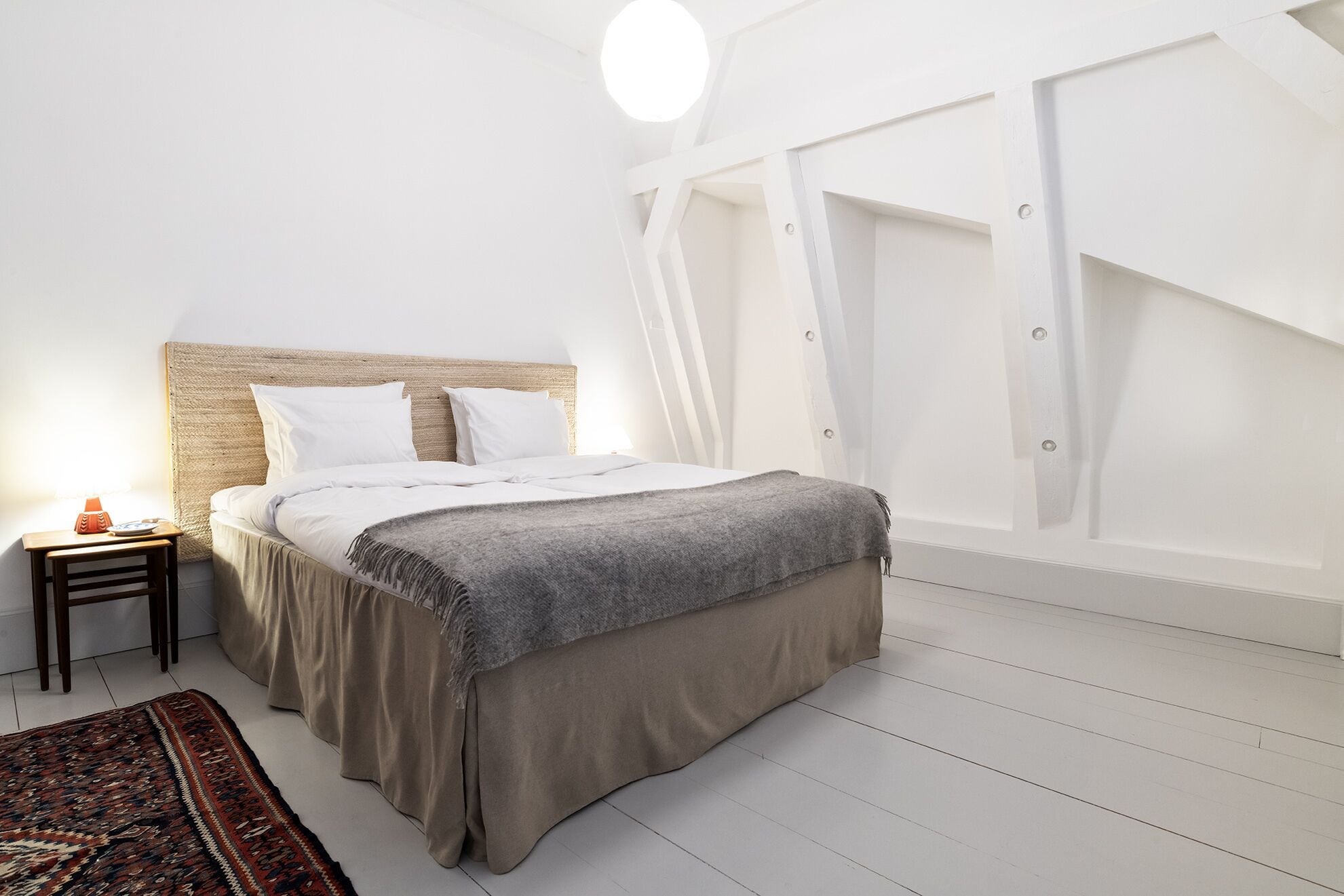 A bedroom with a bed, a bedside table, a rug, and a white wall with a window.