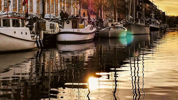 A waterfront with boats docked, buildings with uniform architecture, and a sunset reflecting on the water. - Kanalhuset (Copenhagen)