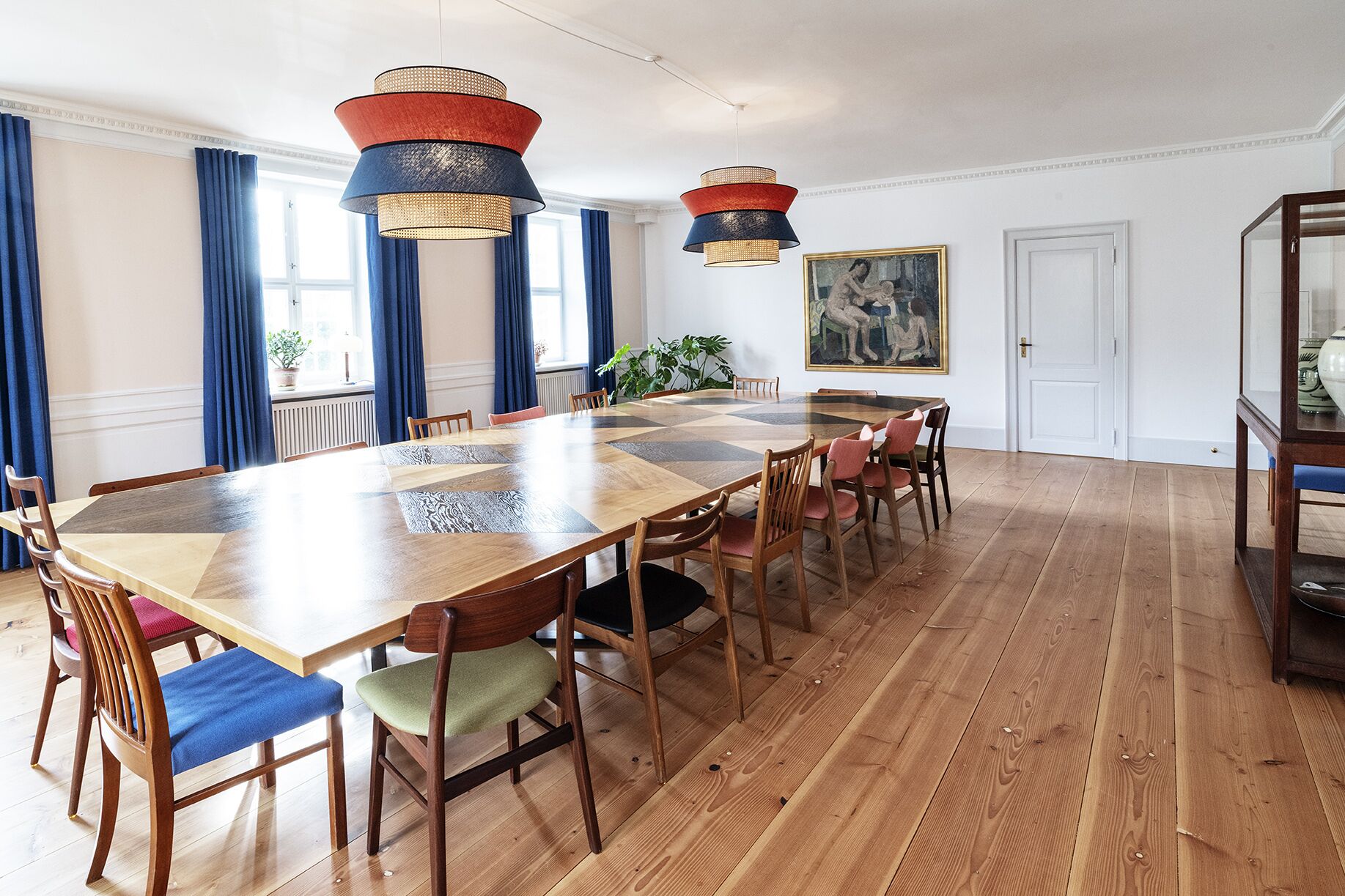 Een ruime eetkamer met een lange houten tafel en stoelen, twee hanglampen en een groot schilderij aan de muur.