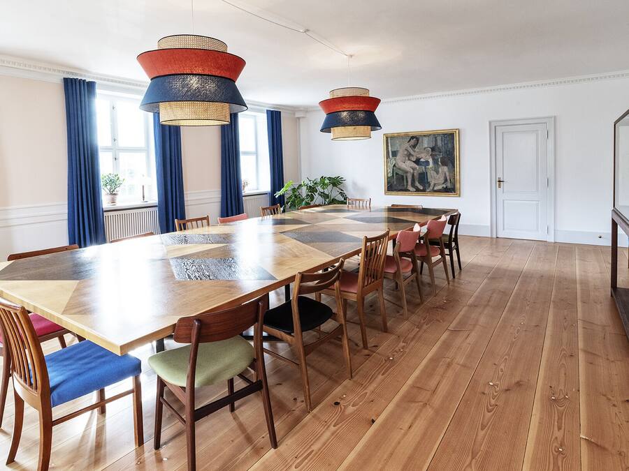 Een ruime eetkamer met een lange houten tafel en stoelen, twee hanglampen en een groot schilderij aan de muur.