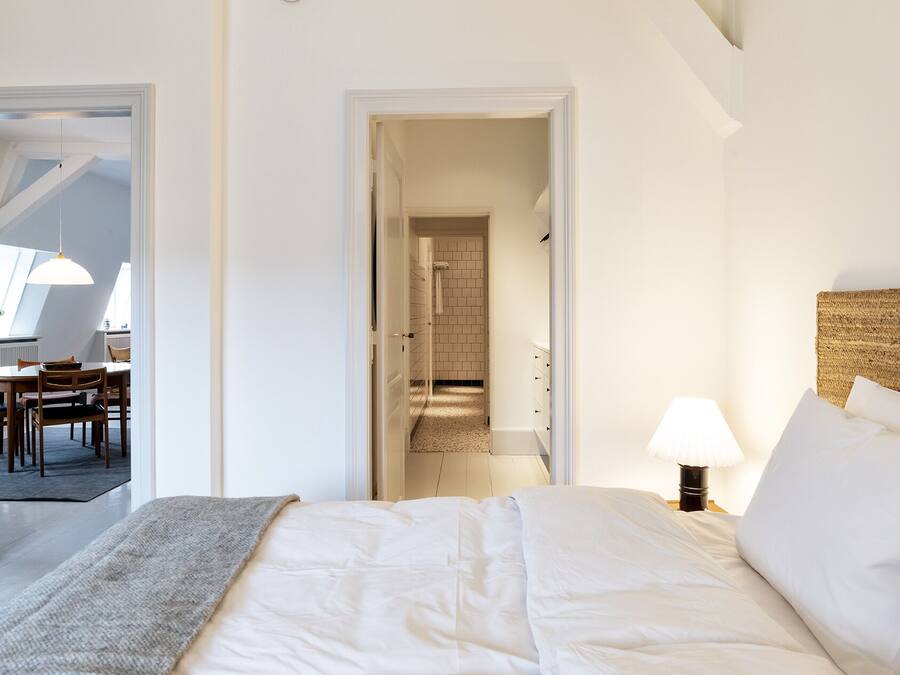 A bedroom with a bed, a nightstand, a lamp, and a view of a dining area through an open door.