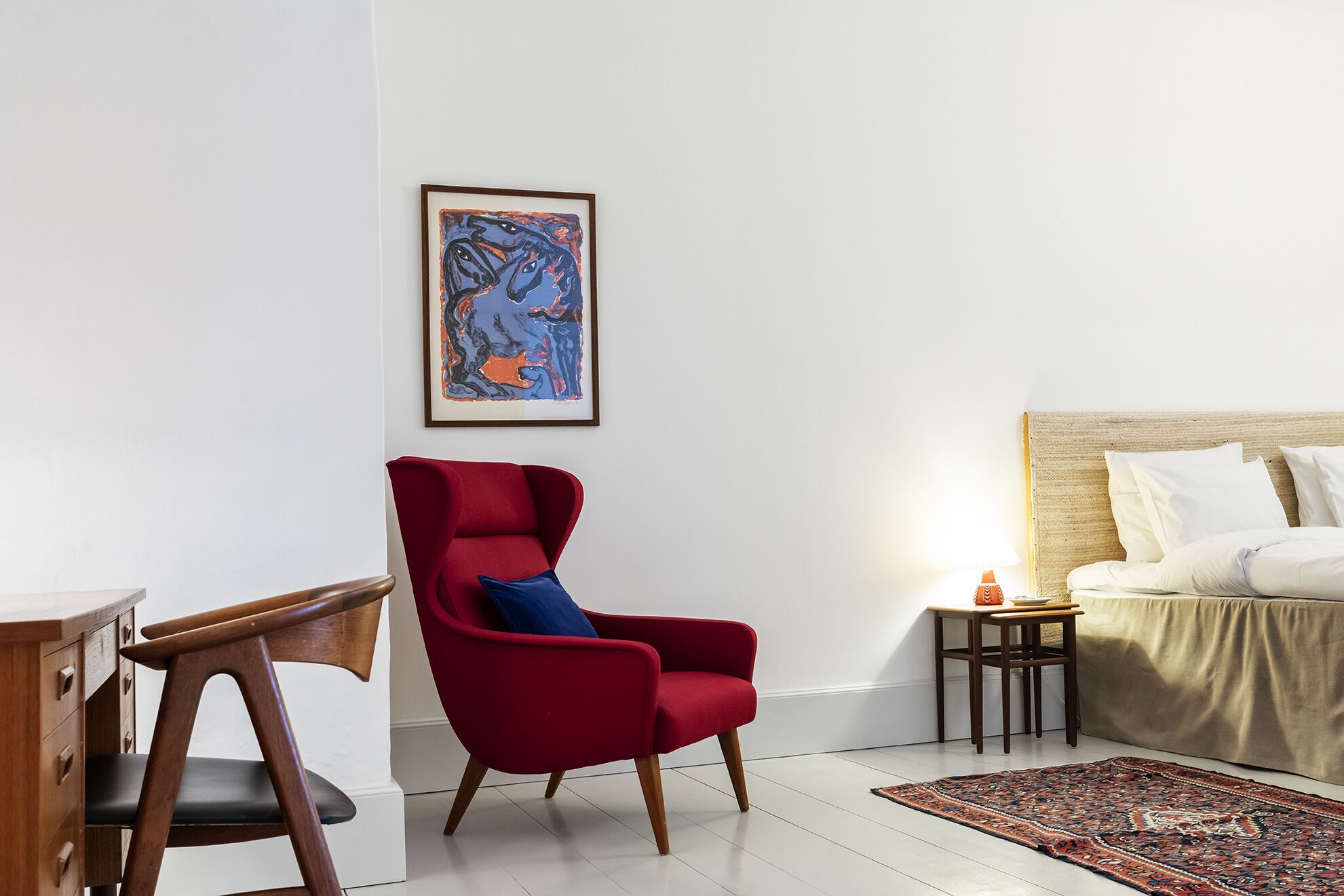 A modern hotel room with a red armchair, a wooden desk, a bed with a beige headboard, and a framed abstract painting on the wall.