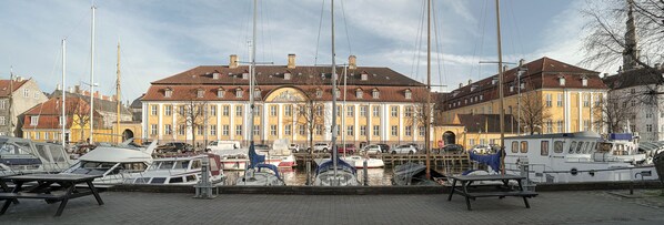 A marina with boats docked, a large yellow building with a red roof, and a clear sky. - Kanalhuset (Copenhagen)