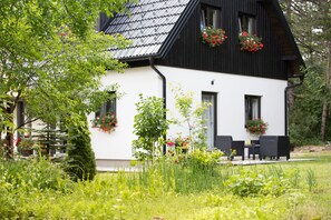 A white house with black trim, surrounded by lush greenery and flowering plants.