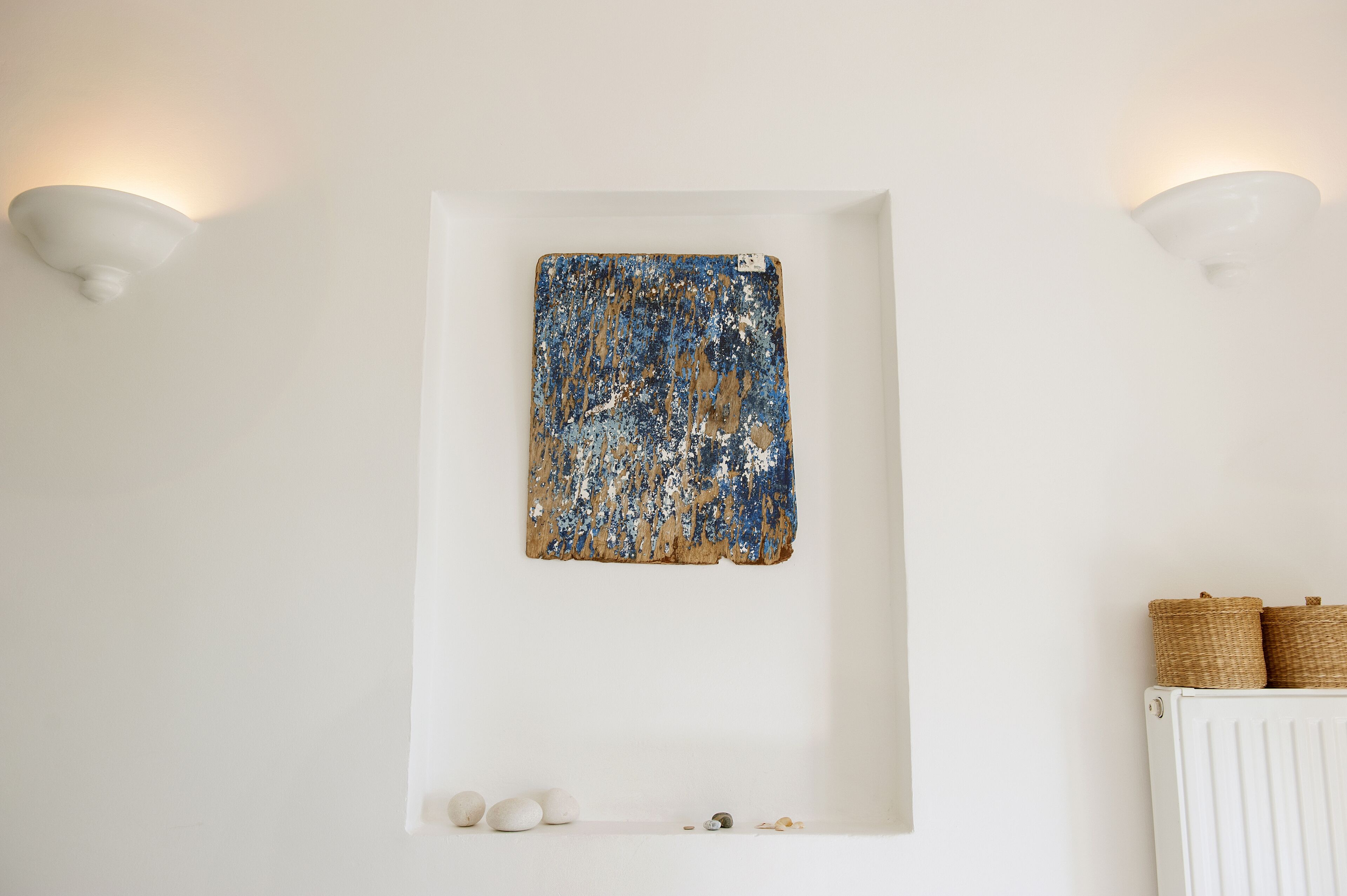 Ein Zimmer mit weißer Wand, einem blau-weißen abstrakten Gemälde, zwei Wandleuchten, einem weißen Heizkörper und einer kleinen Ablage mit Körben.