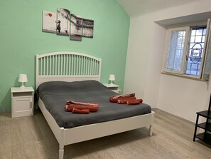 Ein Bett mit weißem Kopfteil, zwei Nachttische mit Lampen, ein Fenster mit Jalousien und ein Kunstwerk an der Wand.