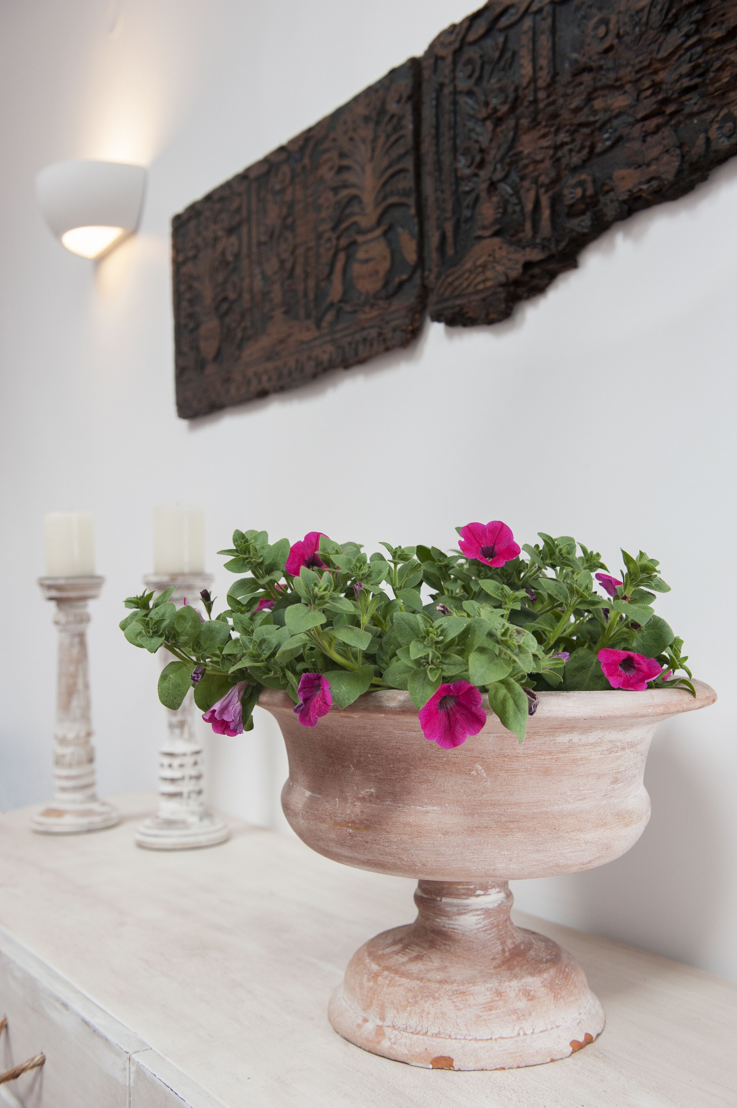 En vit byrå med en keramisk kruka som innehåller rosa blommor, ett vitt ljus och en vit ljusstake.