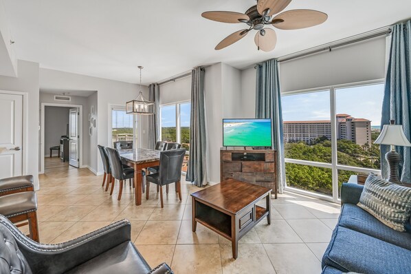 A spacious living room with a dining area, large windows, a ceiling fan, and a TV. - Lockout Unit ~ Private Foyer ~ Community Pool (Miramar Beach)
