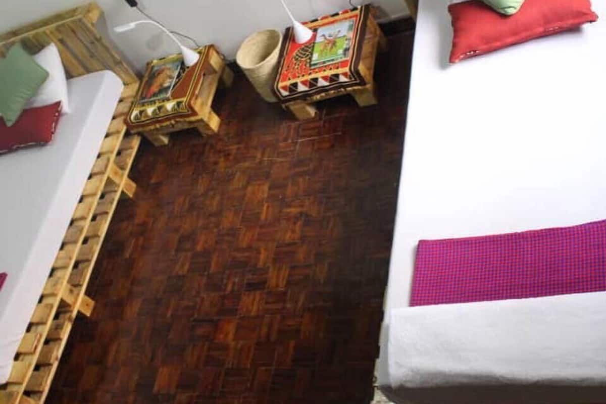 A room with a wooden floor, a bed with a white mattress and a pink blanket, a wooden headboard, a small wooden nightstand with a lamp, and a wall-mounted shelf with decorative items.