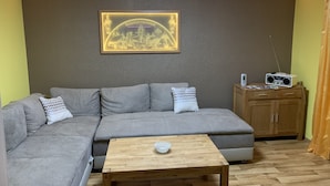 Un salon avec un canapé d’angle gris, une table basse en bois, un tableau encadré au mur, une radio posée sur une commode en bois et un rideau jaune.