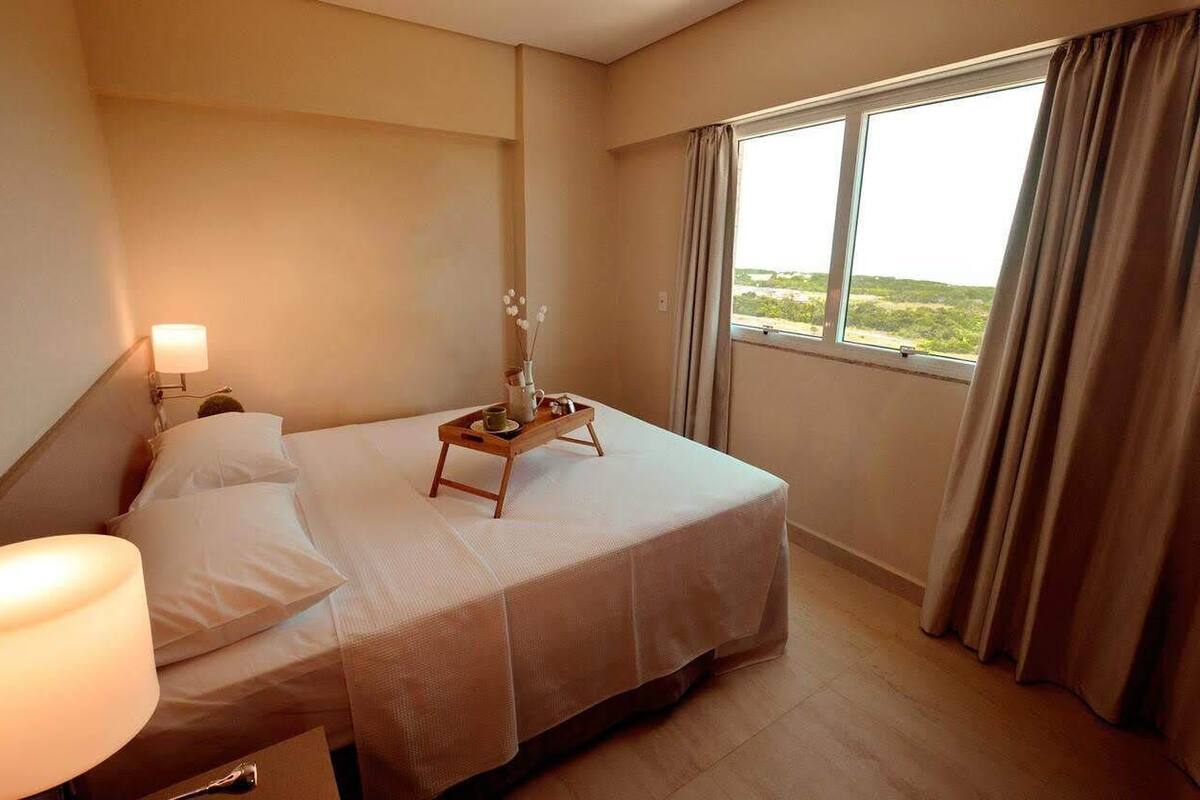 Quarto de hotel com uma cama, uma mesa de cabeceira com abajur, um criado-mudo com um vaso de flores e uma janela com cortinas.