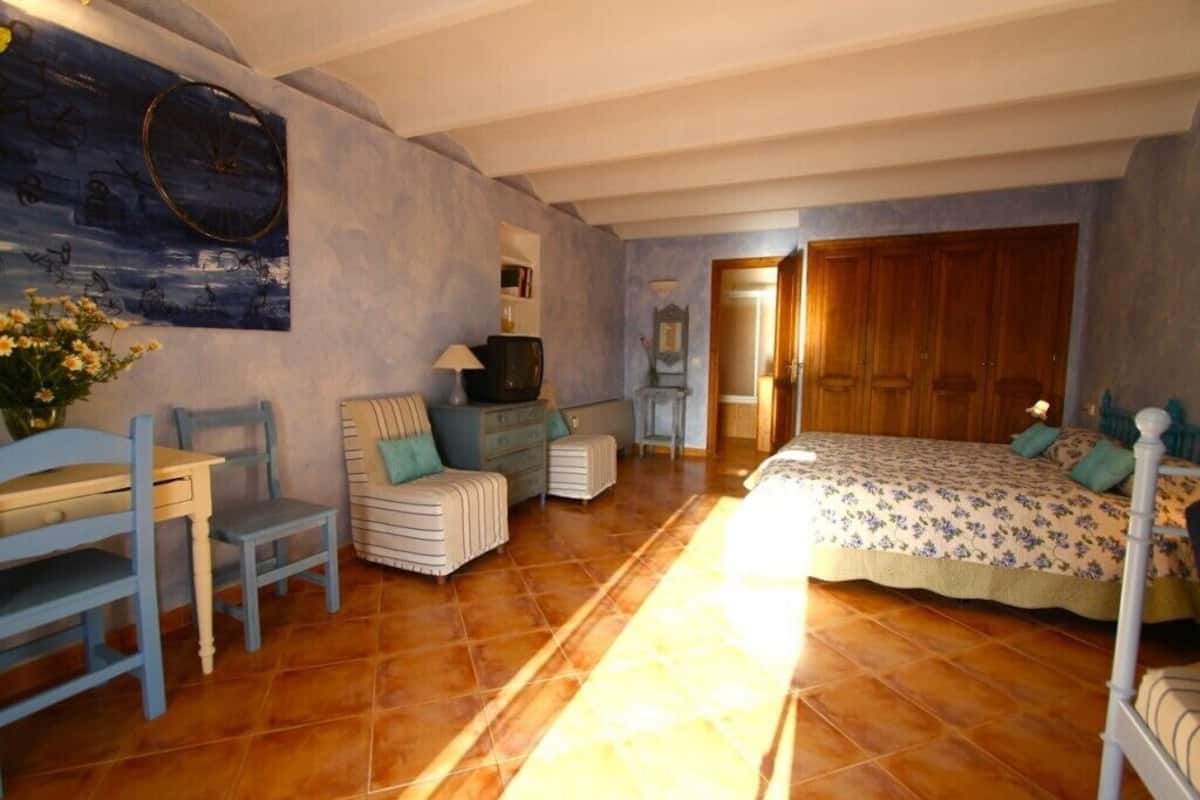 A bedroom with a bed, a chair, a small table, a television, and a painting on the wall.
