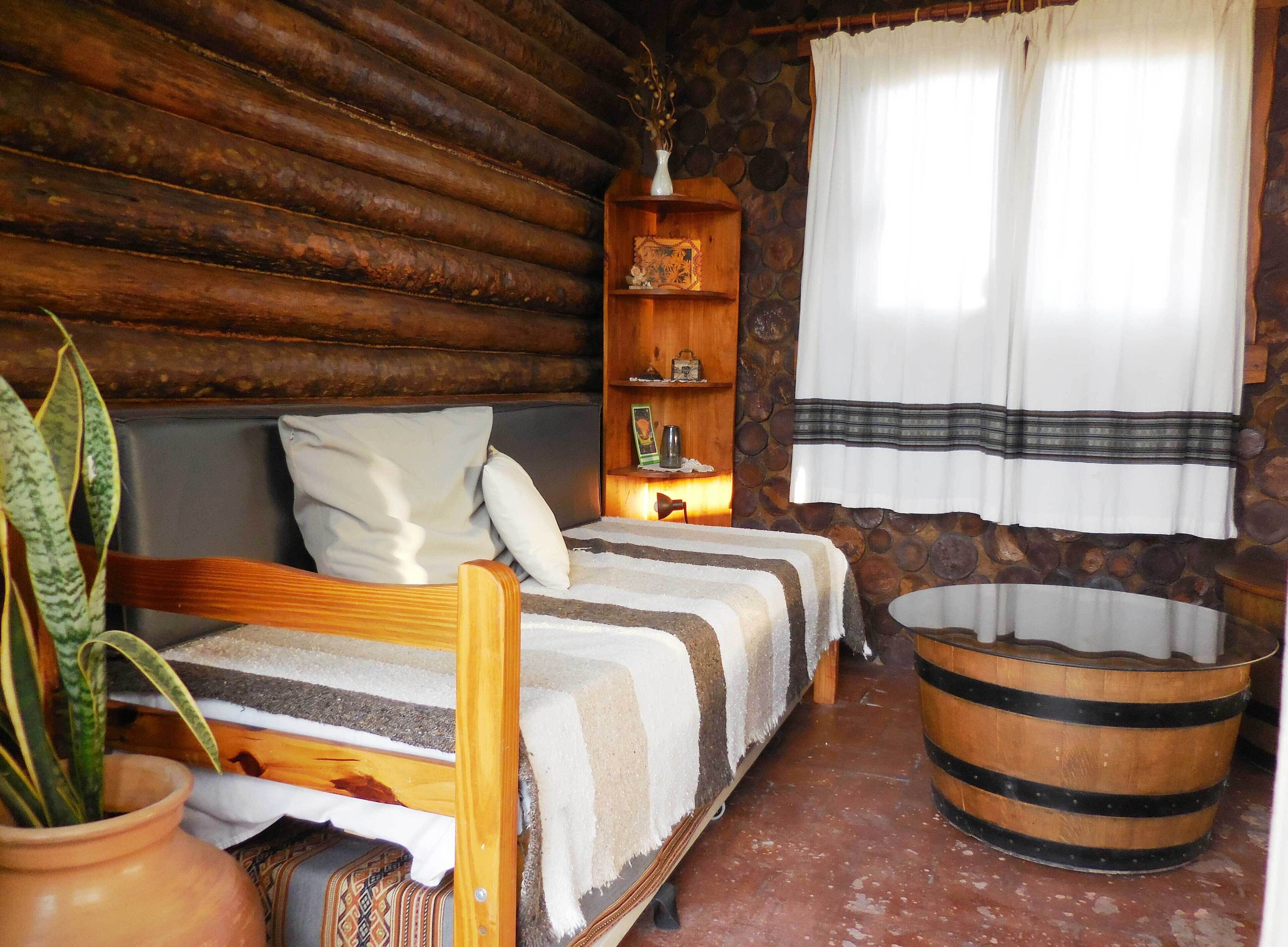 Habitación de cabaña rústica con cama de madera, un pequeño estante con adornos, una cesta grande de fibras naturales y una pared de piedra.