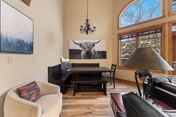 A dining area with a wooden table and benches, a chandelier, and a large window with a view of trees.