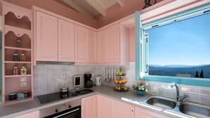 A kitchen with pink cabinets, a stove, a coffee maker, and a window with a view of mountains. - Harmony House (Corfu)