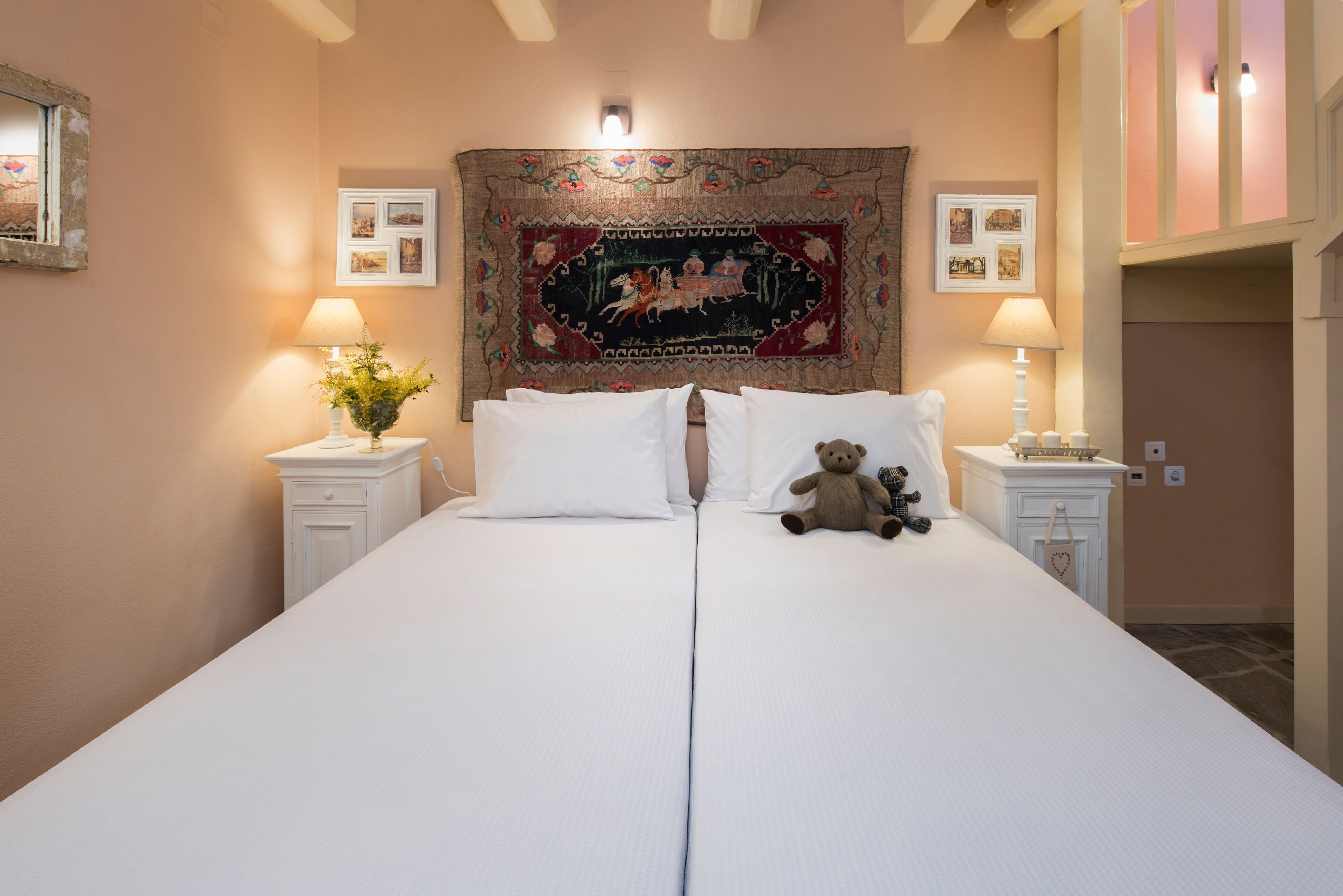 A double bed with white linens, two bedside tables with lamps, a decorative tapestry, and a teddy bear on the bed.