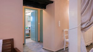 A narrow hallway with a stone floor, a wooden bed, a white chair, and a door leading to a bathroom. - Harmony House (Corfu)