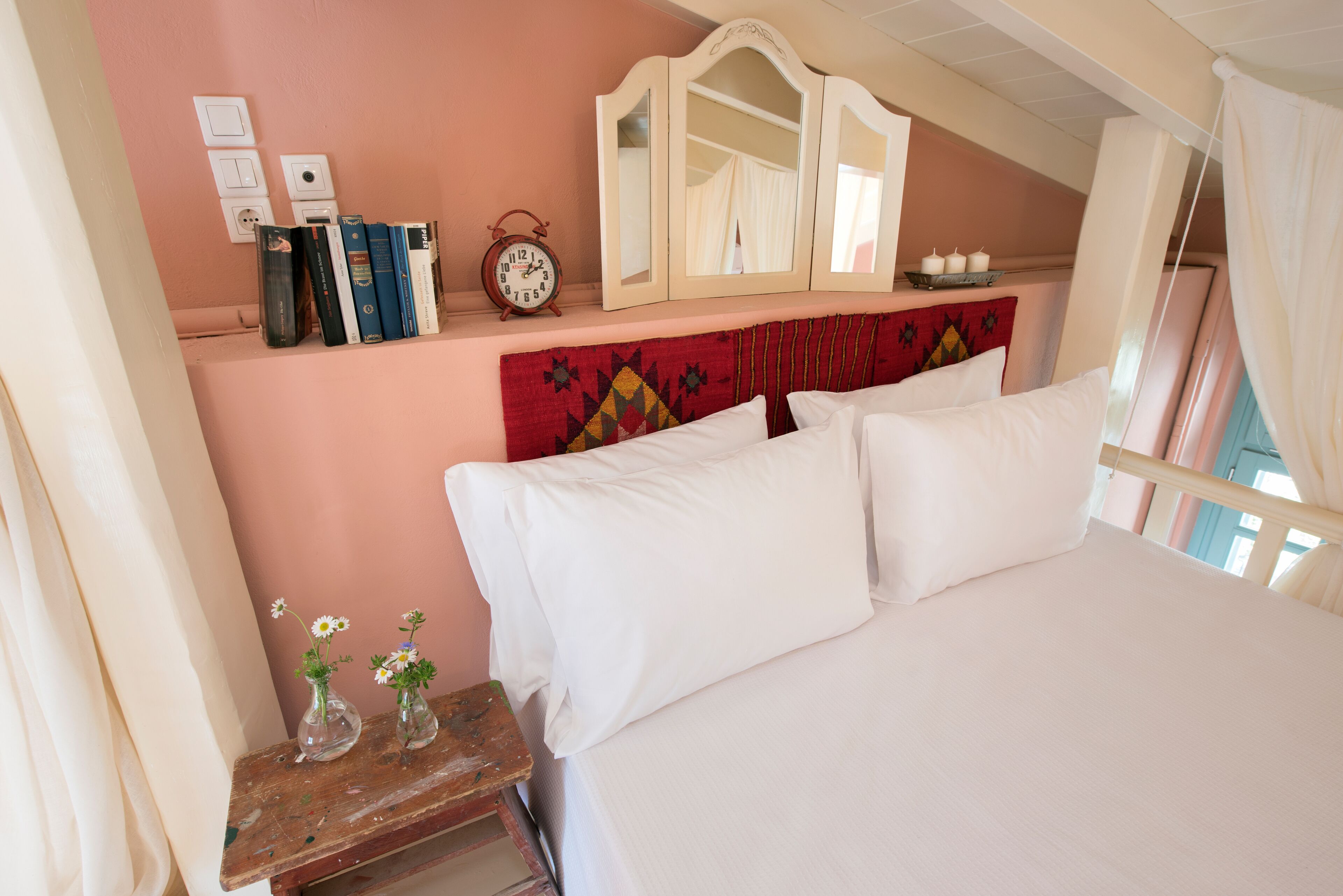 A bedroom with a bed, a bedside table with books and a clock, a mirror, and a wall with light switches.
