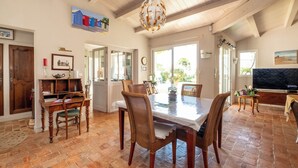 A dining area with a wooden table, wicker chairs, and a chandelier. There is a television on a stand to the right.