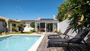 A swimming pool with a wooden deck, lounge chairs, and a glass-enclosed patio area.