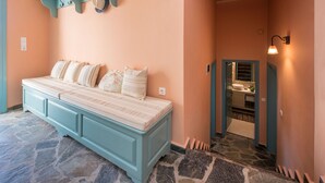 Une chambre avec un grand lit, un sol en pierre, une salle de bain avec un lavabo et un miroir, et un couloir avec un luminaire.