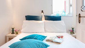 Een net opgemaakt bed met wit en blauw beddengoed, een bedlampje, een boek en een paar zonnebrillen op het bed.