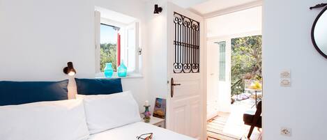 Een slaapkamer met een bed, kussens, een nachtkastje met een lamp, een raam met gordijnen en een deur die naar een buitenruimte leidt.