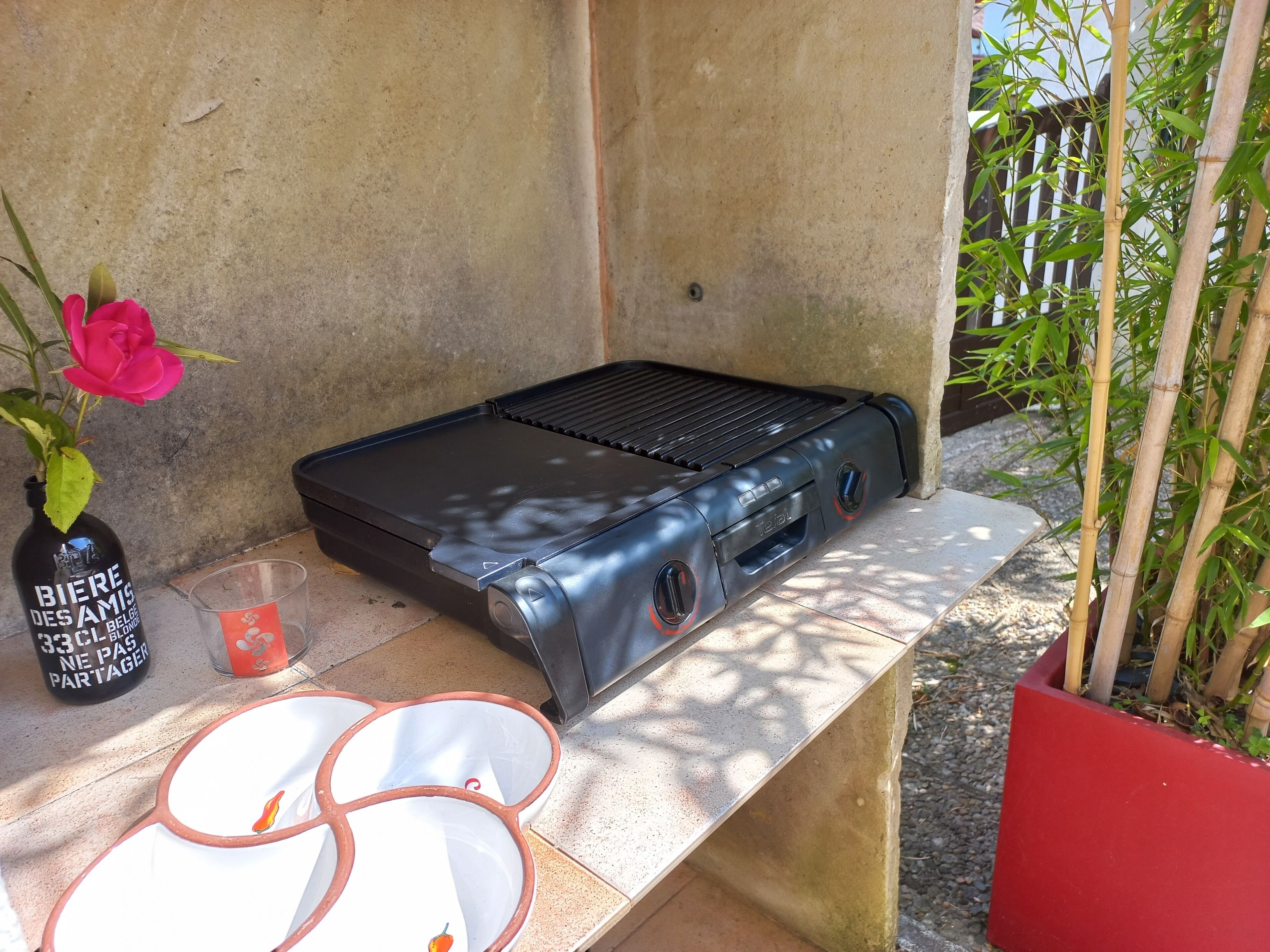 Un barbecue doté d’un panneau de commande, une bouteille de bière, un verre et une fleur rose posés sur une petite étagère en pierre.