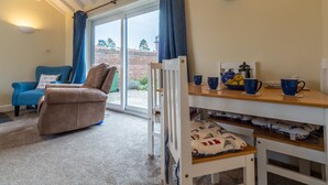 A cozy room with a blue armchair, a brown sofa, a wooden table with cups and a coffee maker, and a white chair with a cushion. - Sea Breeze, Brancaster, Norfolk (Brancaster)