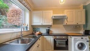 A modern kitchen with a stainless steel sink, a built-in oven, a washing machine, and a window with a view of trees. - Sea Breeze, Brancaster, Norfolk (Brancaster)