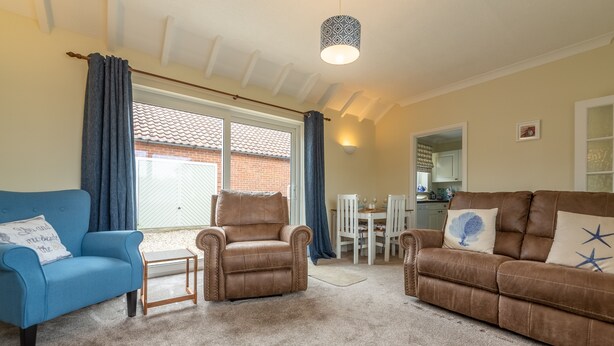 A living room with a blue armchair, two brown leather sofas, a coffee table, and a dining area in the background. - Sea Breeze, Brancaster, Norfolk (Brancaster)