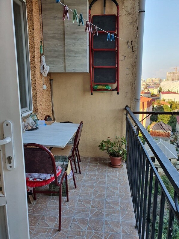 A balcony with a tiled floor, a table with chairs, a potted plant, and a view of buildings in the distance.