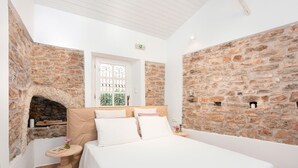 A bedroom with a stone wall, a fireplace, a bed with white bedding, and a small table with a vase. - Nausica House (Corfu)
