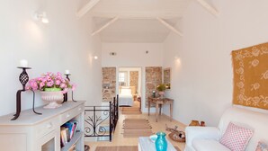 A well-lit room with a white sofa, a coffee table, and a bookshelf. There is a stone wall and a wooden ceiling. - Nausica House (Corfu)