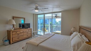 A bedroom with a bed, a TV on a wooden dresser, a ceiling fan, and a balcony with a chair. - Charming Coastal Living: Gulf Views Await (Naples)